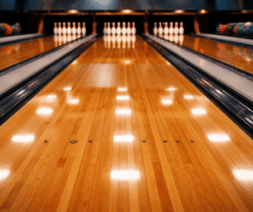 Highly polished wooden bowling alley lanes reflecting overhead lights, with glossy waxed hardwood boards leading toward a full set of white pins at the end of each lane and colorful bowling balls visible along the sides.
